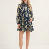 A woman modeling a navy blue long-sleeve mini dress with a beige and gold floral pattern, paired with beige heeled sandals, against a plain white studio background.