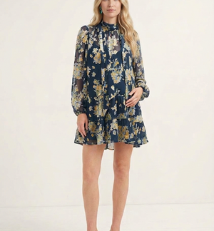 A woman modeling a navy blue long-sleeve mini dress with a beige and gold floral pattern, paired with beige heeled sandals, against a plain white studio background.