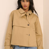 A woman wearing a cropped tan trench-style jacket with a tie collar and front button, paired with light blue jeans, standing against a plain light beige wall.