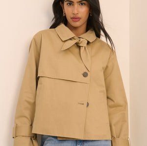A woman wearing a cropped tan trench-style jacket with a tie collar and front button, paired with light blue jeans, standing against a plain light beige wall.
