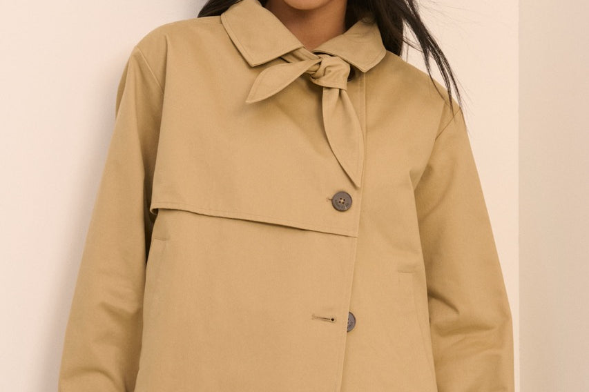 A woman wearing a cropped tan trench-style jacket with a tie collar and front button, paired with light blue jeans, standing against a plain light beige wall.