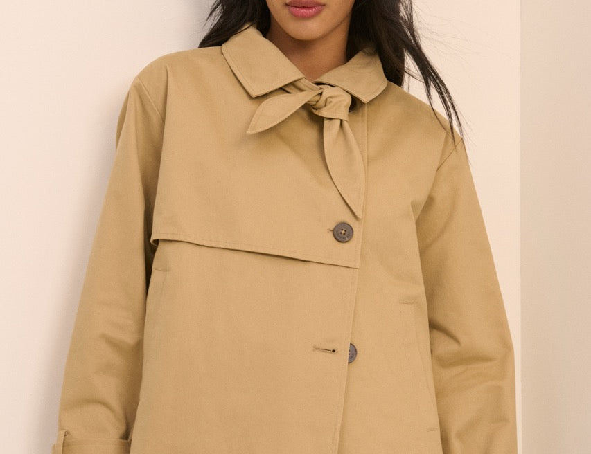 A woman wearing a cropped tan trench-style jacket with a tie collar and front button, paired with light blue jeans, standing against a plain light beige wall.