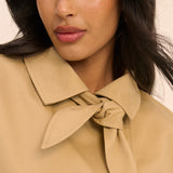 Close-up of a woman wearing a beige or camel-colored blouse with a wide collar and a large front bow tie detail, against a light neutral background.