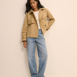 A woman modeling a light tan trench coat over a white top and light blue wide-leg jeans, standing against a plain beige studio background.