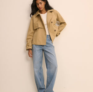 A woman modeling a light tan trench coat over a white top and light blue wide-leg jeans, standing against a plain beige studio background.
