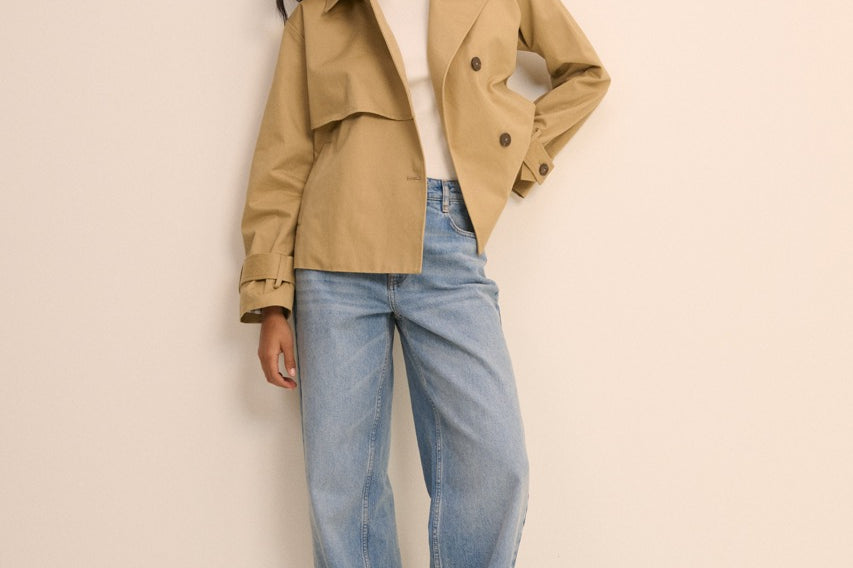 A woman modeling a light tan trench coat over a white top and light blue wide-leg jeans, standing against a plain beige studio background.