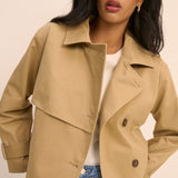 A woman wearing a light tan double-breasted trench jacket over a white top and blue jeans, photographed against a plain beige studio background.