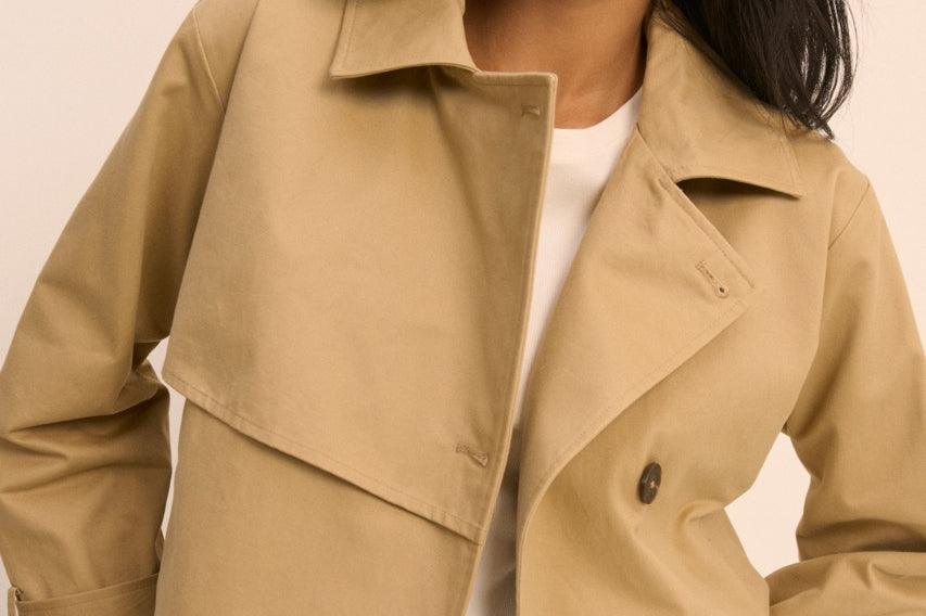 A woman wearing a light tan double-breasted trench jacket over a white top and blue jeans, photographed against a plain beige studio background.