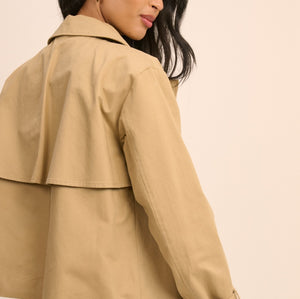 A woman wearing a cropped tan trench jacket with rolled cuffs and blue jeans, shown from a side-back angle against a plain beige background.