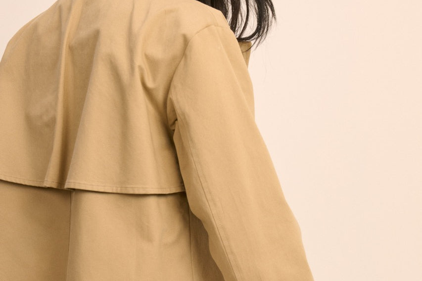 A woman wearing a cropped tan trench jacket with rolled cuffs and blue jeans, shown from a side-back angle against a plain beige background.