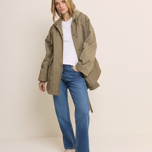 A woman modeling an oversized olive green hooded jacket over a white top and blue jeans, standing against a plain light beige studio background.