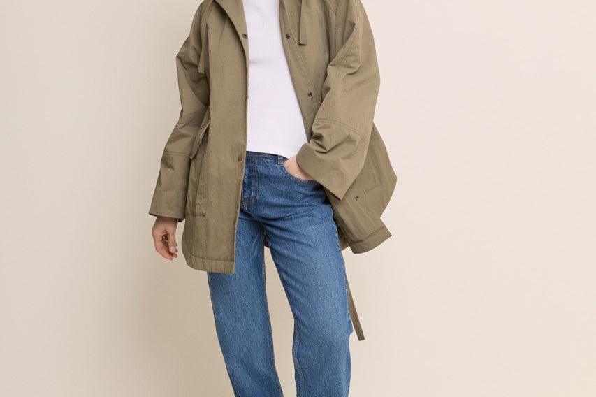 A woman modeling an oversized olive green hooded jacket over a white top and blue jeans, standing against a plain light beige studio background.