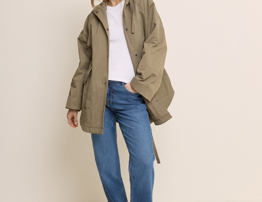 A woman modeling an oversized olive green hooded jacket over a white top and blue jeans, standing against a plain light beige studio background.