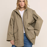 A woman modeling an oversized olive green hooded jacket with front flap pockets, worn over a white t-shirt and blue jeans, against a solid beige background.