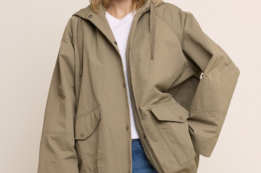 A woman modeling an oversized olive green hooded jacket with front flap pockets, worn over a white t-shirt and blue jeans, against a solid beige background.