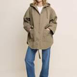 A woman modeling an oversized olive green hooded jacket with a drawstring, worn over a white top and blue jeans, standing against a plain light beige studio background.