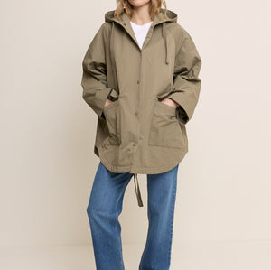 A woman modeling an oversized olive green hooded jacket with a drawstring, worn over a white top and blue jeans, standing against a plain light beige studio background.