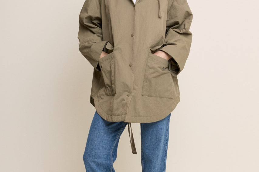 A woman modeling an oversized olive green hooded jacket with a drawstring, worn over a white top and blue jeans, standing against a plain light beige studio background.