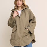 A woman modeling an oversized olive green hooded jacket with front pockets, worn over a white top and blue jeans, against a plain beige studio background.