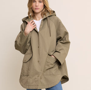 A woman modeling an oversized olive green hooded jacket with front pockets, worn over a white top and blue jeans, against a plain beige studio background.
