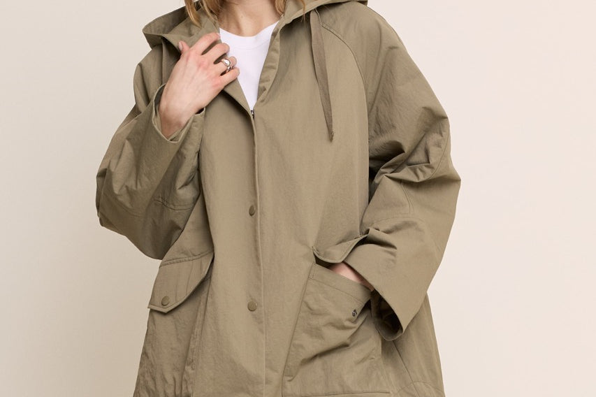 A woman modeling an oversized olive green hooded jacket with front pockets, worn over a white top and blue jeans, against a plain beige studio background.