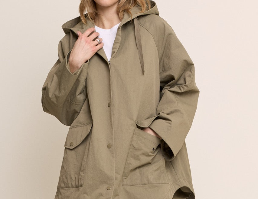 A woman modeling an oversized olive green hooded jacket with front pockets, worn over a white top and blue jeans, against a plain beige studio background.