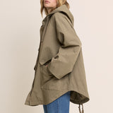 A woman wearing an oversized olive green hooded jacket with large front patch pockets and drawstrings, paired with blue jeans, photographed against a plain light beige studio background.