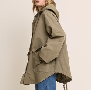 A woman wearing an oversized olive green hooded jacket with large front patch pockets and drawstrings, paired with blue jeans, photographed against a plain light beige studio background.