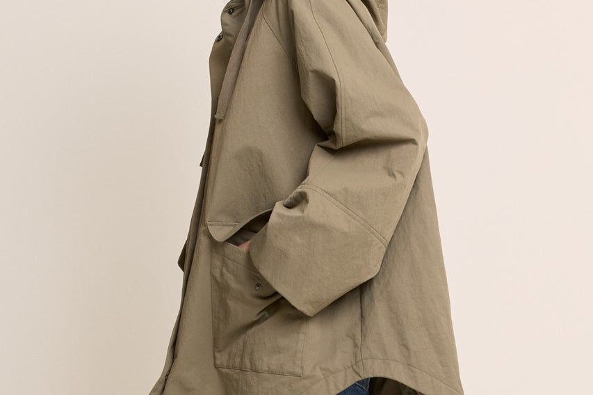 A woman wearing an oversized olive green hooded jacket with large front patch pockets and drawstrings, paired with blue jeans, photographed against a plain light beige studio background.