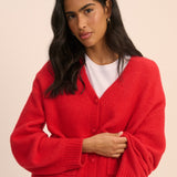 A woman modeling a bright red knit cardigan sweater with a white t-shirt and blue jeans, posed against a solid light beige background.