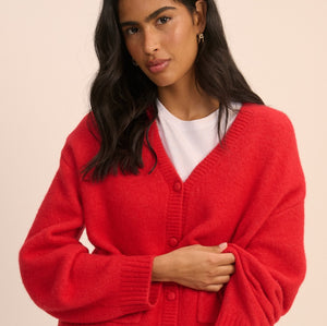 A woman modeling a bright red knit cardigan sweater with a white t-shirt and blue jeans, posed against a solid light beige background.