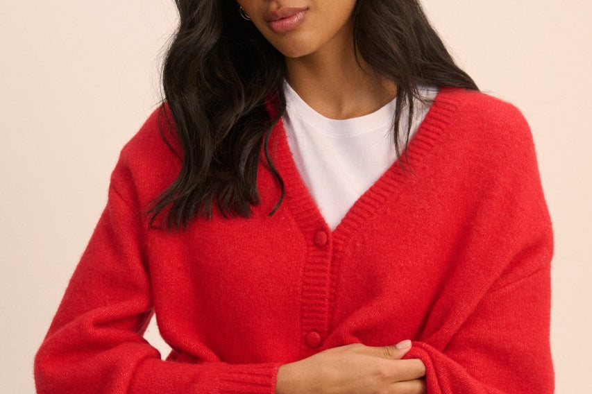 A woman modeling a bright red knit cardigan sweater with a white t-shirt and blue jeans, posed against a solid light beige background.