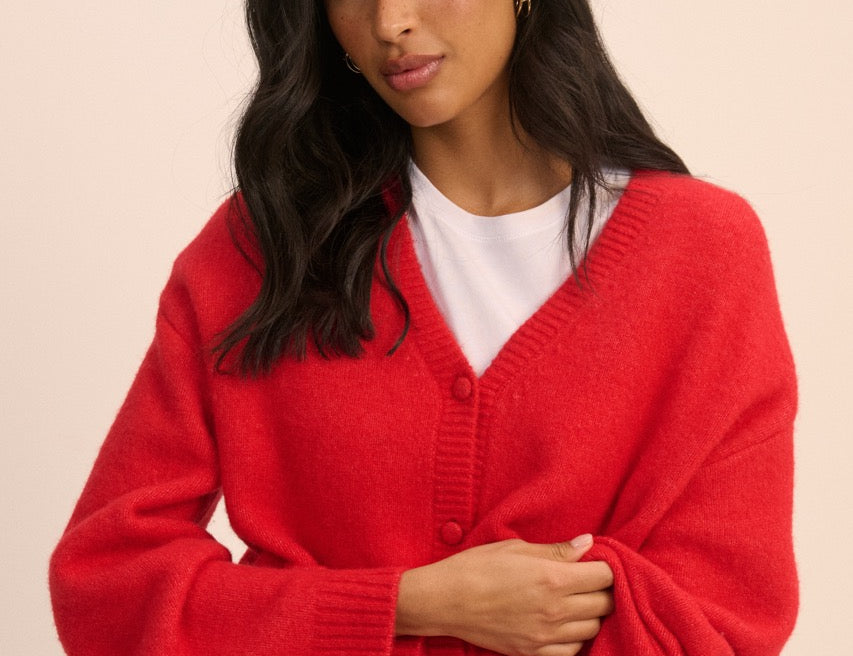 A woman modeling a bright red knit cardigan sweater with a white t-shirt and blue jeans, posed against a solid light beige background.