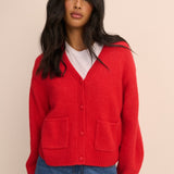 A woman wearing a bright red knit cardigan with a V-neck, front buttons, and two large patch pockets, layered over a white t-shirt and blue jeans, against a solid beige background.