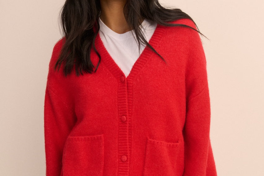 A woman wearing a bright red knit cardigan with a V-neck, front buttons, and two large patch pockets, layered over a white t-shirt and blue jeans, against a solid beige background.