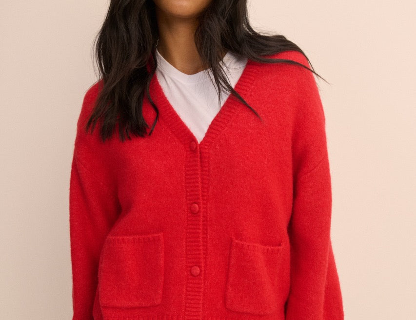 A woman wearing a bright red knit cardigan with a V-neck, front buttons, and two large patch pockets, layered over a white t-shirt and blue jeans, against a solid beige background.