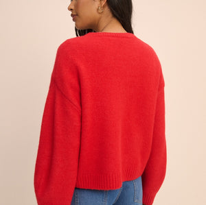 A woman shown from the back wearing a bright red knit sweater and blue jeans against a plain beige studio background.