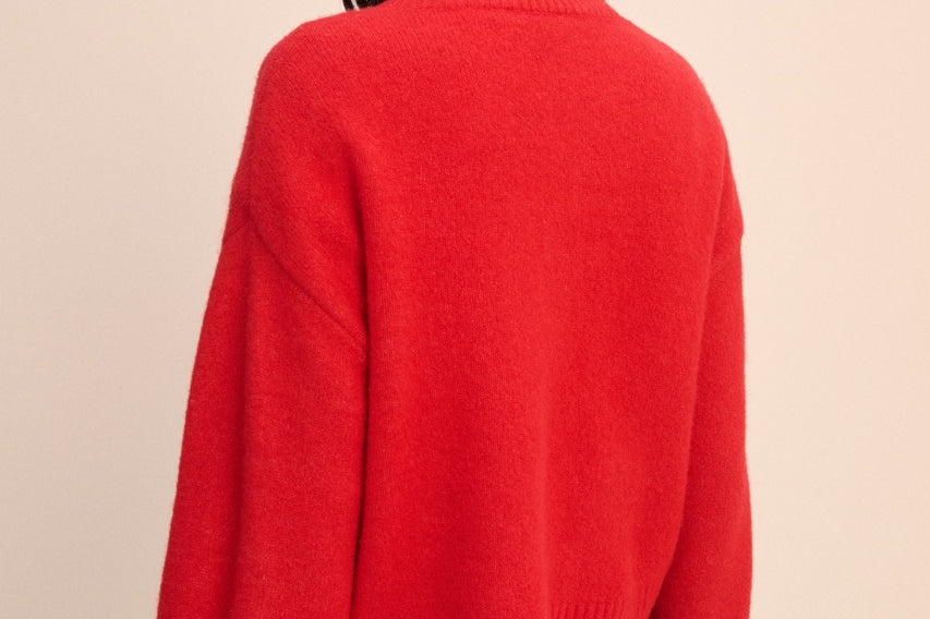 A woman shown from the back wearing a bright red knit sweater and blue jeans against a plain beige studio background.