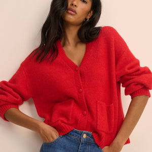 A woman wearing a bright red knit cardigan sweater with a V-neck and front buttons, paired with blue jeans, posing against a solid light beige background.