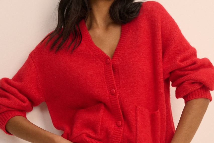A woman wearing a bright red knit cardigan sweater with a V-neck and front buttons, paired with blue jeans, posing against a solid light beige background.