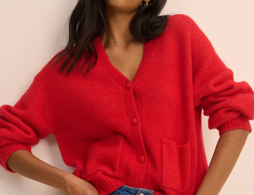 A woman wearing a bright red knit cardigan sweater with a V-neck and front buttons, paired with blue jeans, posing against a solid light beige background.