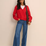 A woman modeling a bright red cardigan sweater, blue wide-leg jeans, and brown shoes, with a brown shoulder bag, against a solid light beige background.