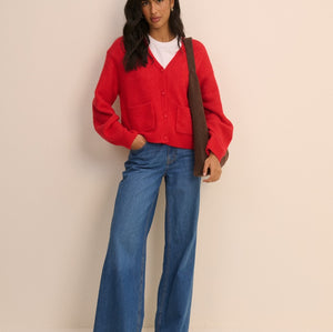 A woman modeling a bright red cardigan sweater, blue wide-leg jeans, and brown shoes, with a brown shoulder bag, against a solid light beige background.