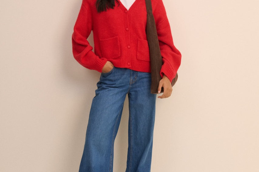 A woman modeling a bright red cardigan sweater, blue wide-leg jeans, and brown shoes, with a brown shoulder bag, against a solid light beige background.