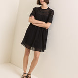 A woman modeling a short black dress with sheer mesh sleeves and a gathered waist, standing in a minimalist cream-colored room corner.