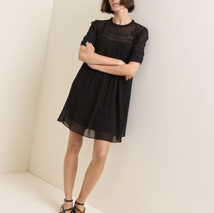 A woman modeling a short black dress with sheer mesh sleeves and a gathered waist, standing in a minimalist cream-colored room corner.
