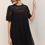 A woman wearing a black short-sleeve pleated dress with sheer mesh sleeves and a lace yoke, posing against a plain beige wall.
