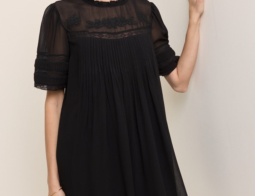 A woman wearing a black short-sleeve pleated dress with sheer mesh sleeves and a lace yoke, posing against a plain beige wall.