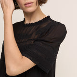 A close-up of a woman wearing a black sheer short-sleeve blouse with lace trim and a ruffled neckline, posed against a light beige background.