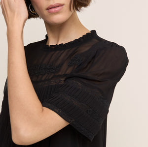 A close-up of a woman wearing a black sheer short-sleeve blouse with lace trim and a ruffled neckline, posed against a light beige background.